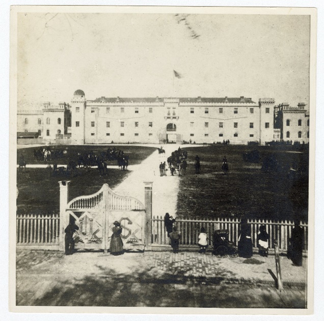 The original Citadel building on Marion Square, Charleston, seen through its front gate, with a flag above the building and onlookers along the fence